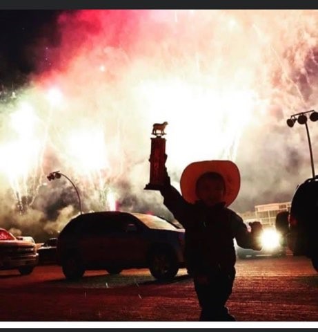 A young cowboy in a cowboy hat holds up a trophy with fireworks in the background - Discover Surrey Through My Lens